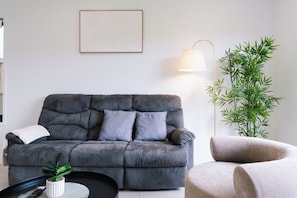 Ein modernes Wohnzimmer mit einem grauen Sofa, einem kleinen runden Tisch mit einer Pflanze, einer Lampe und einem gerahmten Bild an der Wand.