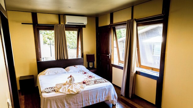 Una habitación de hotel con cama, mesita de noche, unidad de aire acondicionado y ventanas grandes con cortinas.