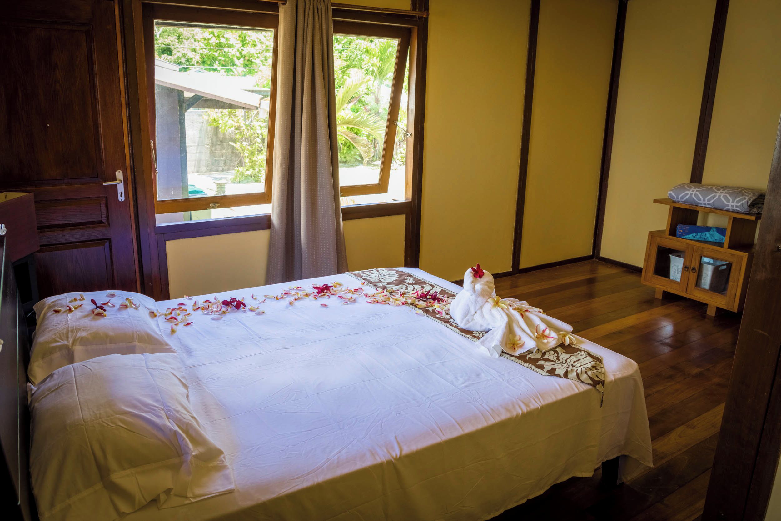 A hotel room with a bed, wooden floor, a window with curtains, and a small wooden shelf with items.