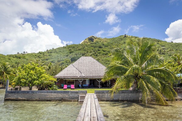 A thatched-roof bungalow with a wooden dock leading to a clear lagoon, surrounded by lush greenery and a hill in the background.