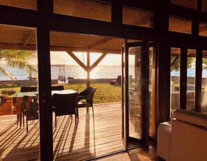 Una terraza de madera con una mesa y sillas, con vista a una playa con palmeras y una puesta de sol.