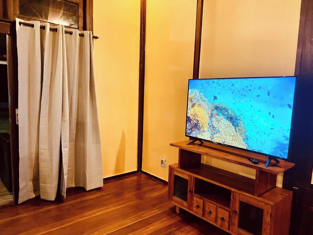 A room with a wooden floor, a television on a wooden stand, and a window with curtains.