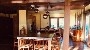 Un comedor amplio con una larga mesa de madera y sillas, una cocina al fondo y ventanas grandes que dejan entrar luz natural.
