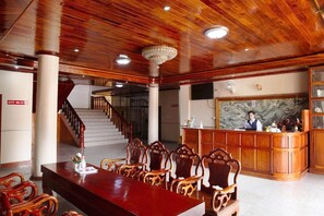 A hotel lobby with a reception desk, a staircase, and a dining area with wooden chairs and a long table. - Mittaphap Ii Hotel (Luang Prabang)