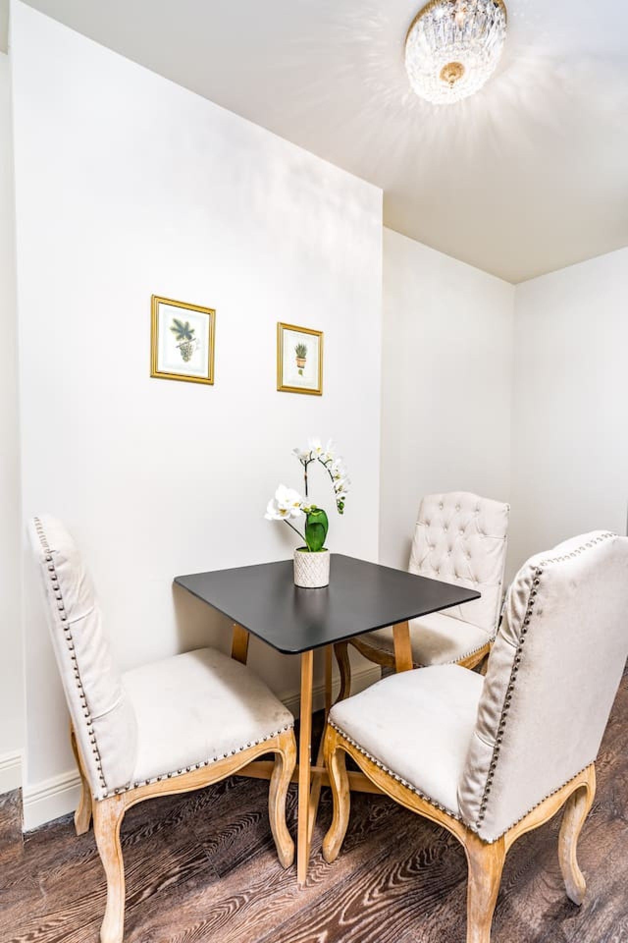 A corner room with a dark wooden table, two upholstered chairs with decorative stitching, and two framed botanical prints on the wall.