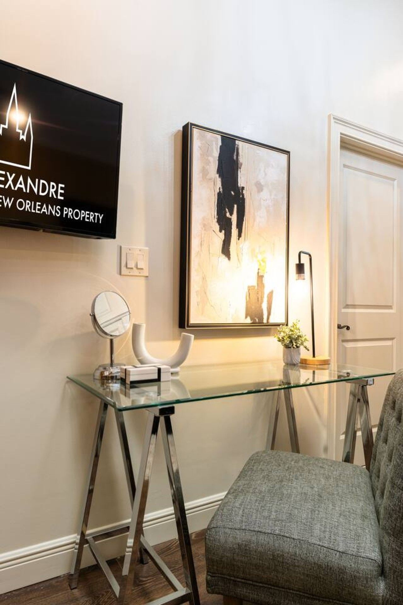 A modern interior with a glass desk, a framed abstract painting, and a television displaying 'XANDRE NEW ORLEANS PROPERTY'.