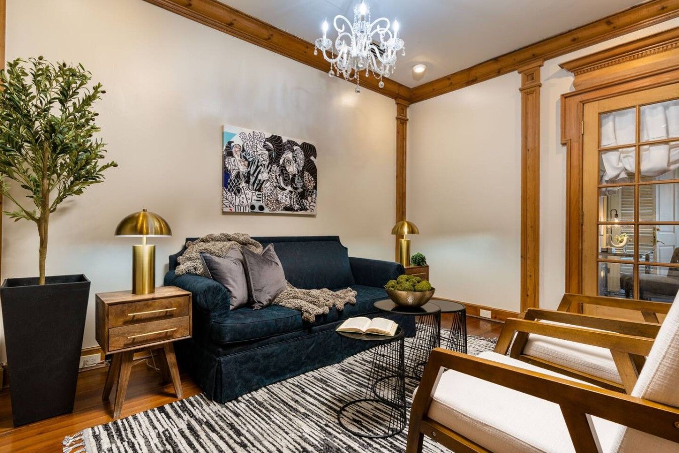 Un salon chaleureux avec un canapé bleu foncé, une table d’appoint en bois, un lampadaire, une plante en pot et une grande fenêtre avec des rideaux.