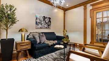 Un salon chaleureux avec un canapé bleu foncé, une table d’appoint en bois, un lampadaire, une plante en pot et une grande fenêtre avec des rideaux.