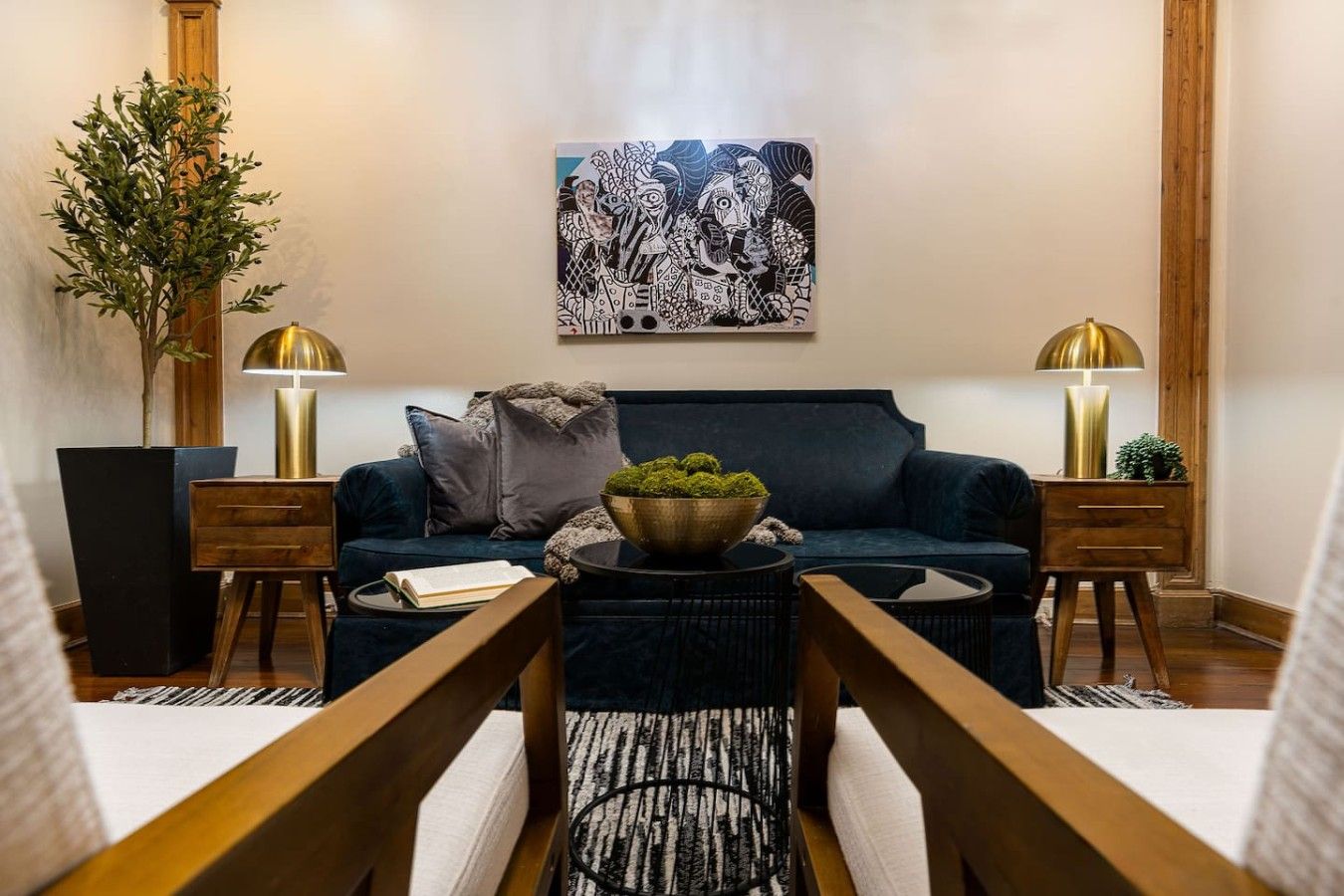 Un salon moderne avec un canapé bleu foncé, deux tables d’appoint en bois, une lampe sur pied, une table basse agrémentée d’une plante verte et une œuvre d’art abstraite accrochée au mur.
