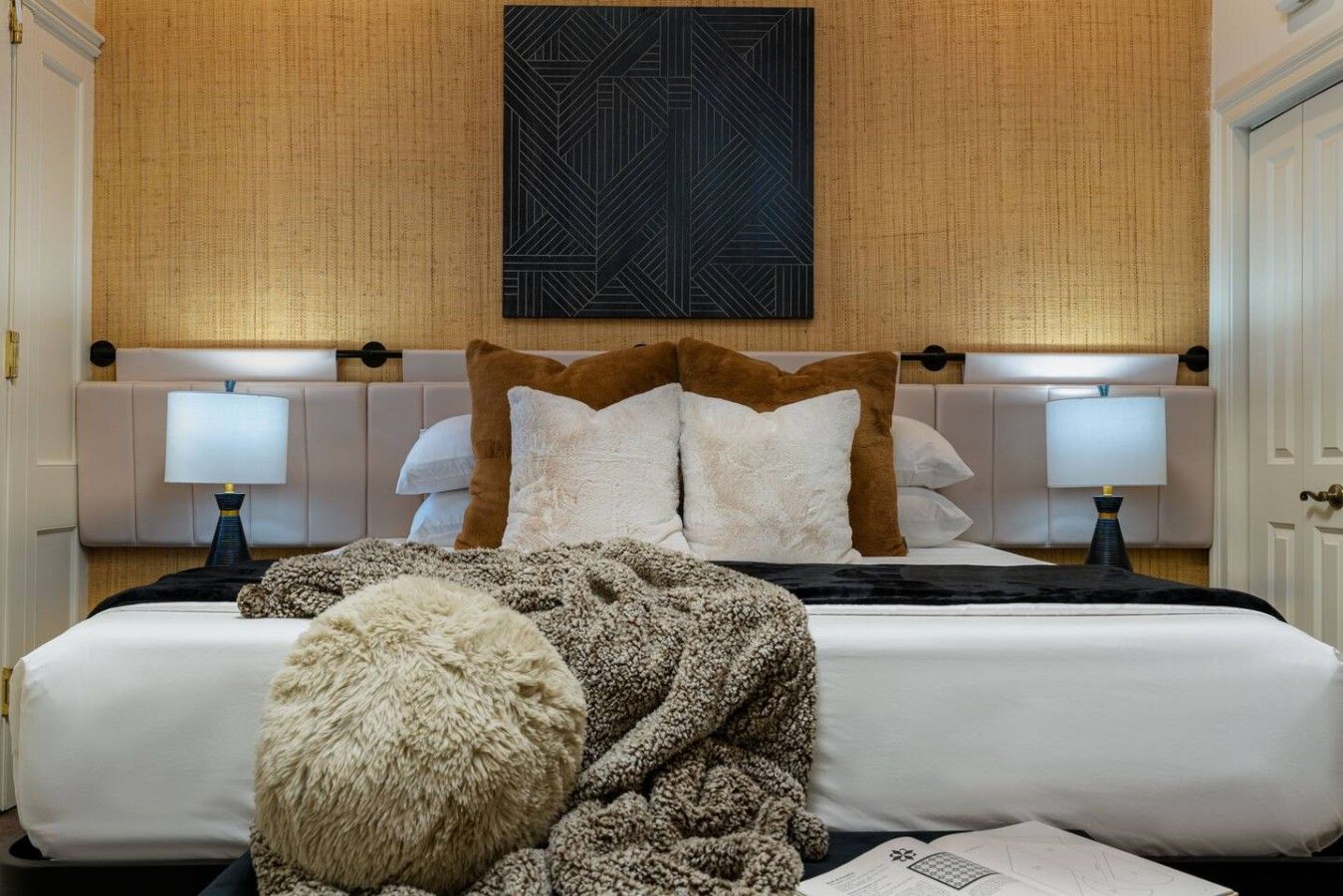 A hotel room with a large bed, two bedside tables with lamps, a headboard with a geometric pattern, and a fluffy throw blanket on the bed.