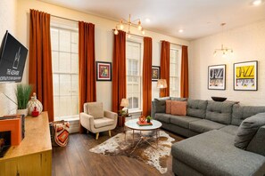 A living room with a sofa, armchair, and a coffee table. There are curtains, a wall-mounted TV, and framed artwork. - Modern 2BD in CBD | Walk to Bourbon St (New Orleans)