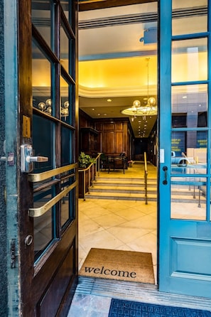 A hotel entrance with a wooden door, a 'welcome' mat, and a staircase leading to a lobby area. - Bourbon Bliss | 2BD CBD Condo Near Canal St (New Orleans)