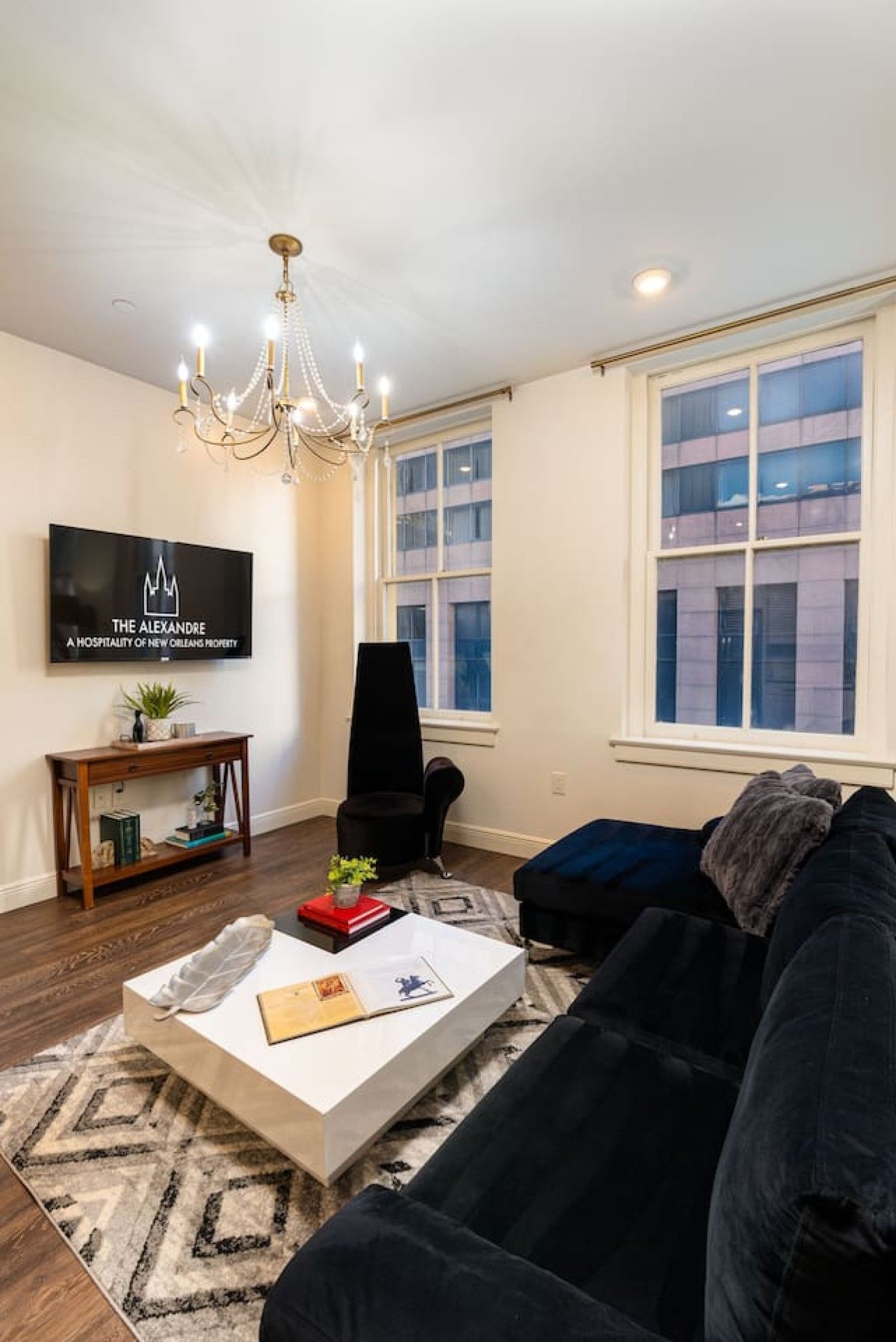 A living room with a black sofa, a white coffee table, a black chair, a TV on the wall, and a chandelier.