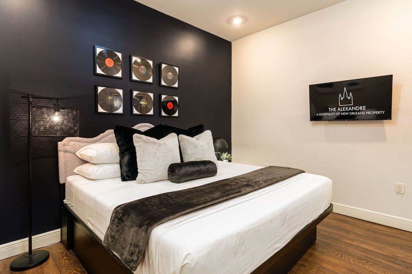 A modern hotel room with a large bed, a wall-mounted TV, and a series of vinyl records displayed above the bed.
