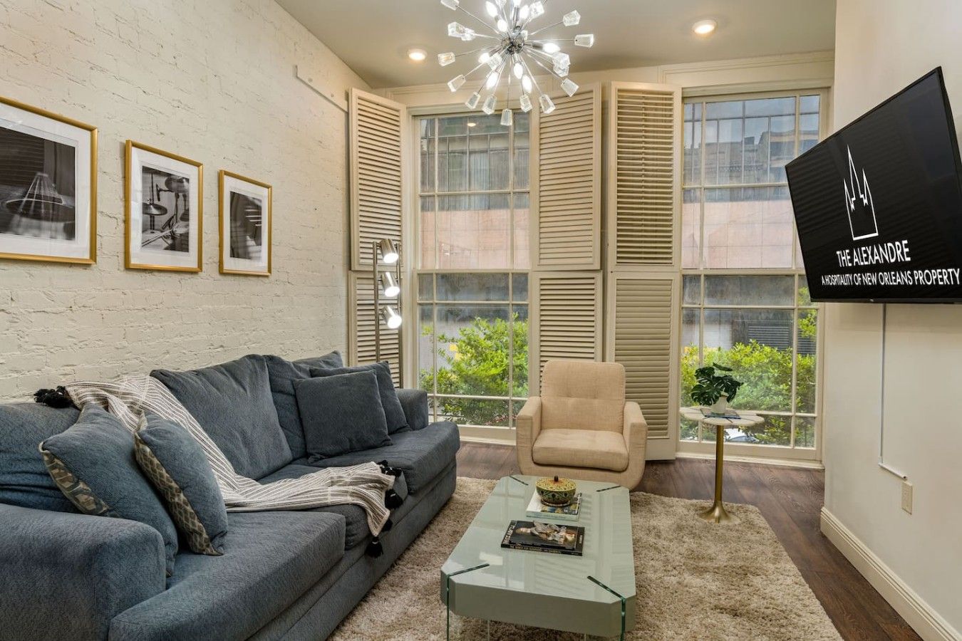 A modern living room with a grey sofa, a beige armchair, and a glass coffee table. There are framed pictures on the wall, a chandelier, and a sign for 'The Alexandre.'