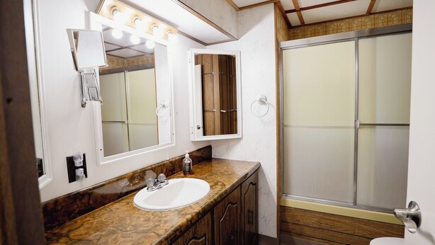A bathroom with a marble countertop, a white sink, a mirror with lights, a wooden cabinet, and a shower with a frosted glass door. - River Cabin is in Cascade right on the Missouri River (Cascade)