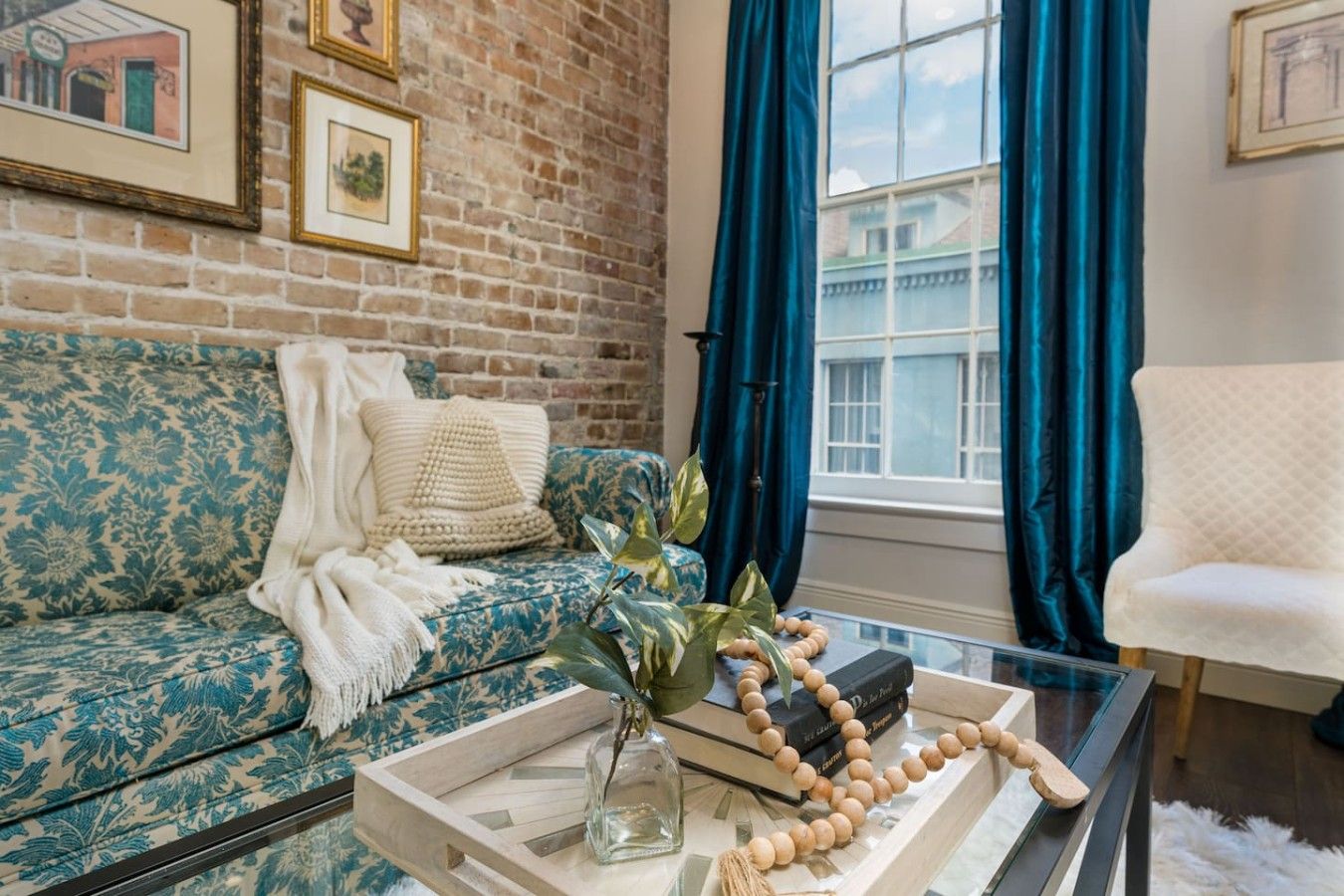 A living room with a patterned sofa, a glass coffee table with decorative items, a brick wall, and a window with curtains.