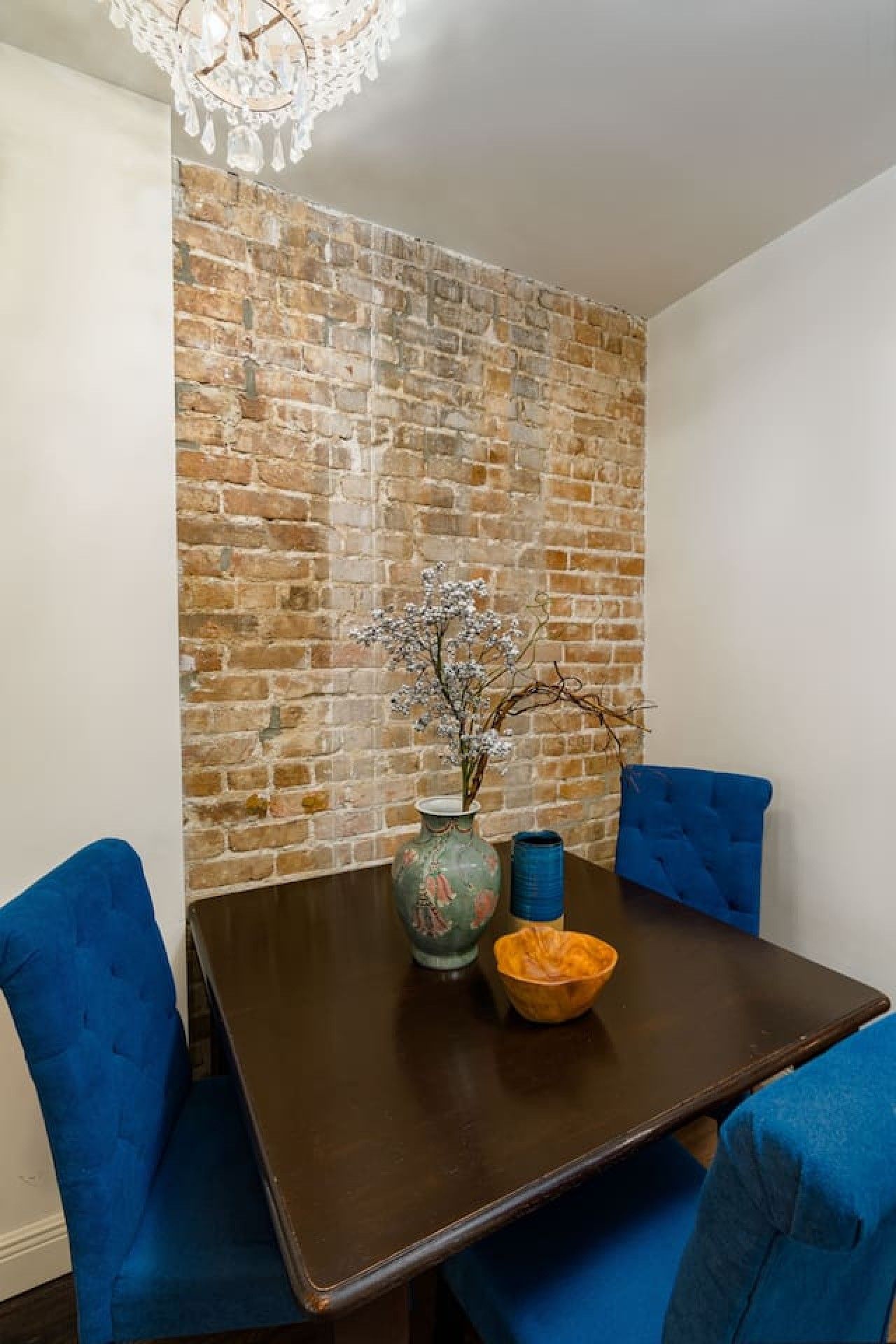 Un coin repas avec un mur en briques, un vase de fleurs et des chaises recouvertes d’un tissu bleu.