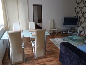 Un salon moderne avec une table à manger en verre, des chaises blanches, un canapé, un téléviseur et un papier peint à motifs.