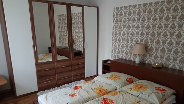 Un dormitorio con un armario de madera, una cama con ropa de cama estampada, una mesita de noche y un papel tapiz decorado.