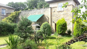 A stone building with a green awning, surrounded by a well-maintained garden with various shrubs and a lawn.