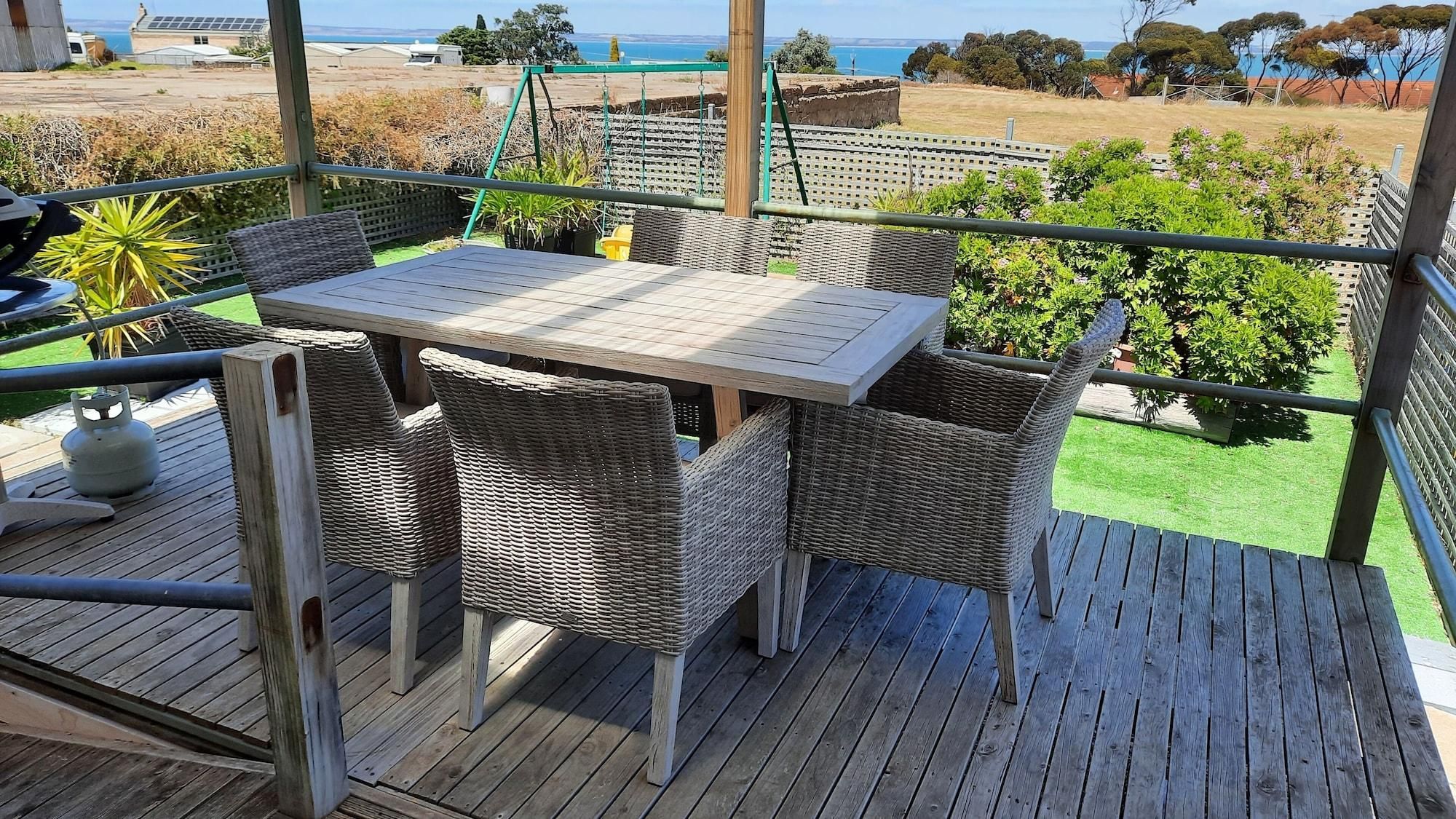 Una terraza de madera con una mesa y sillas de mimbre, con vistas a un área verde con árboles y una vista distante del mar.