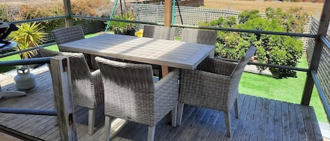 A wooden deck with a table and wicker chairs, overlooking a grassy area with trees and a distant view of the sea.