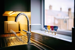 A kitchen sink with a modern faucet, a dish rack, and colorful glassware on a windowsill.