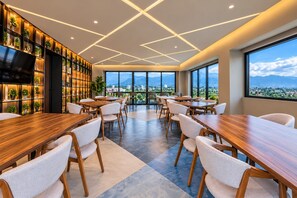 Un comedor moderno con mesas y sillas de madera, una pared de plantas en macetas y vistas a la ciudad y montañas a través de amplios ventanales.