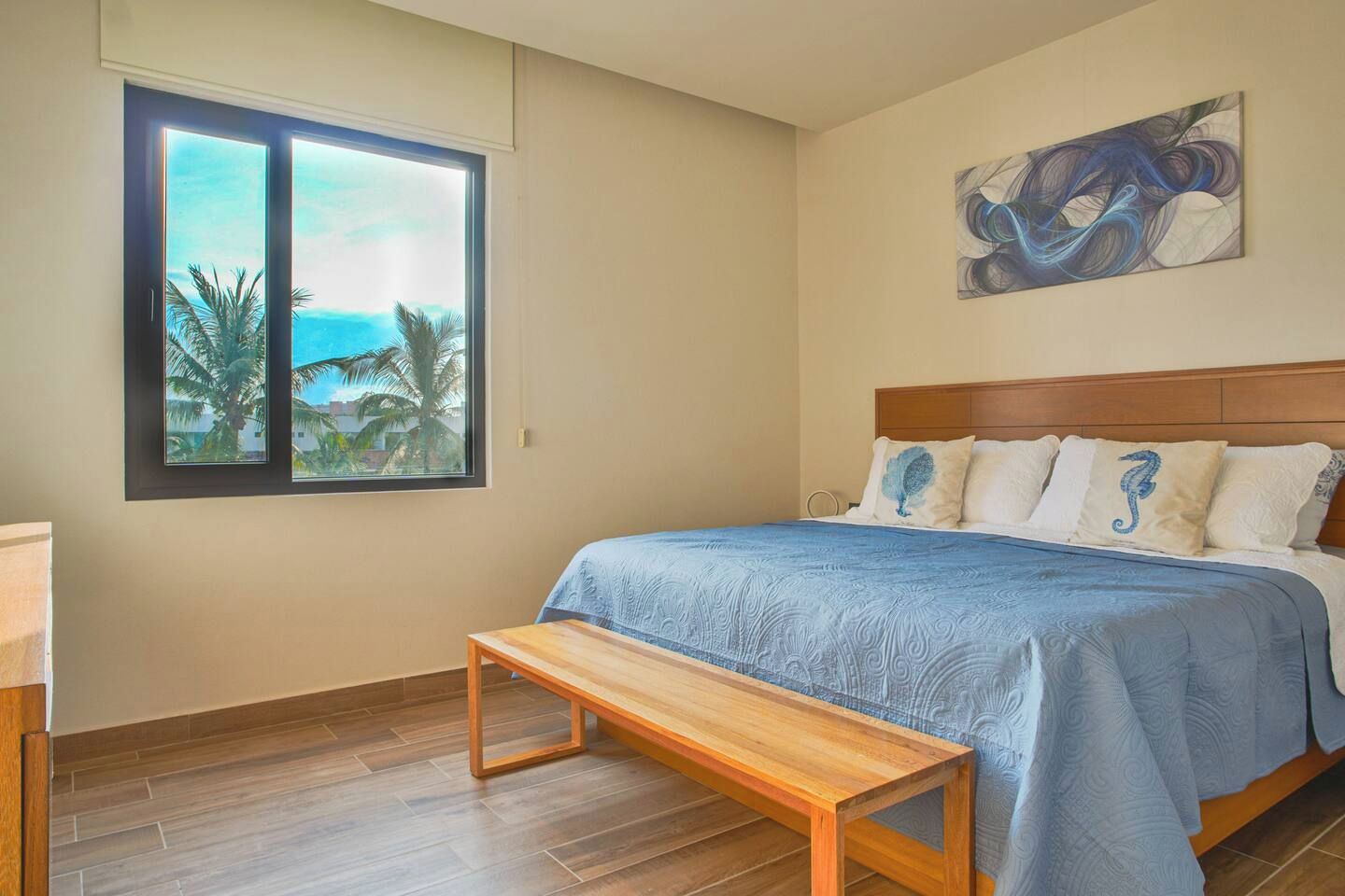 A bedroom with a bed, a bench, a window with a view of palm trees, and a painting on the wall.