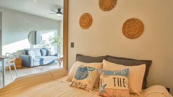 A modern bedroom with a large bed, two wall-mounted decorative items, and a view of a living area with a sofa and a dining table.