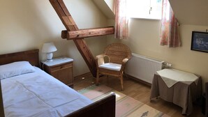 Ein Zimmer mit schrägen Wänden, einem Bett, einem Sessel, einem Tisch und einem fenster mit Vorhängen.