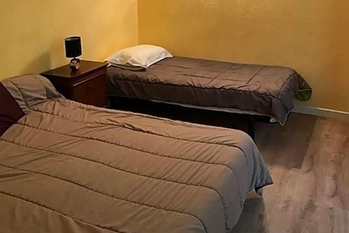 Quarto com duas camas, uma mesa de cabeceira e luminária fixada na parede.