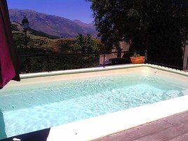 Piscina retangular com água cristalina, rodeada por um deck de madeira e um guarda-corpo metálico. Ao fundo, montanhas e um céu limpo.