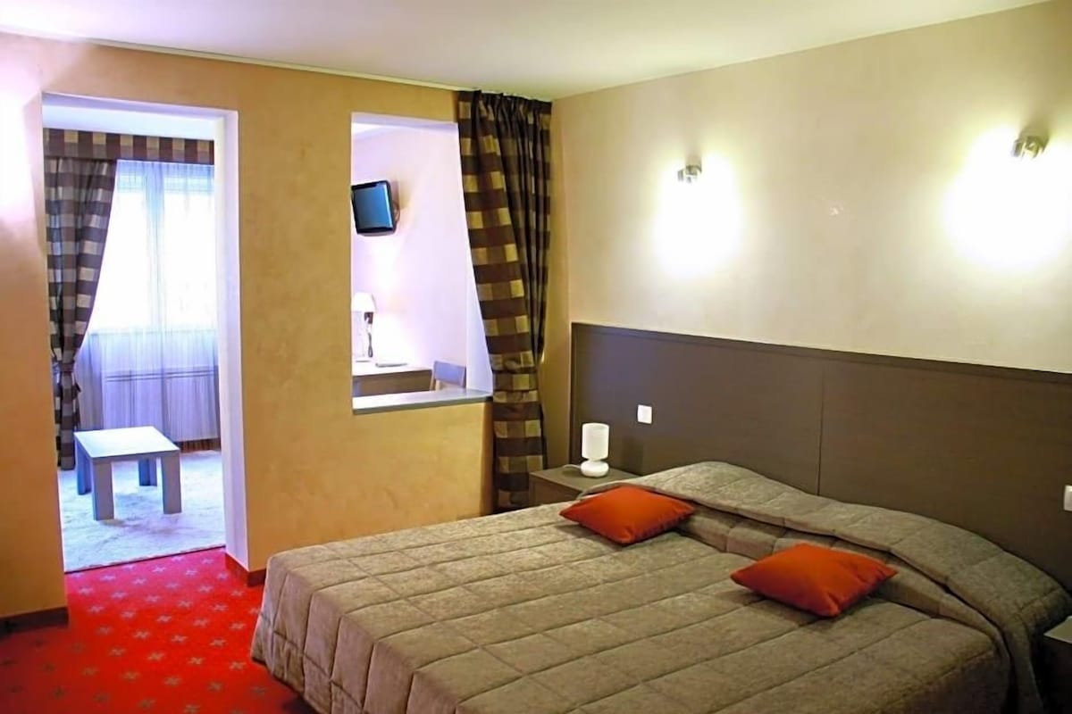 Quarto de hotel com cama, mesa de cabeceira, televisão e janela com cortinas.