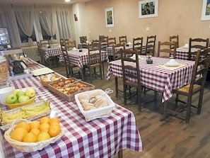 Uma área de refeições com várias mesas cobertas por toalhas xadrez, cadeiras de madeira e cestos de frutas e doces.