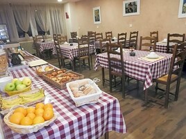 Uma área de refeições com várias mesas cobertas por toalhas xadrez, cadeiras de madeira e cestos de frutas e doces.