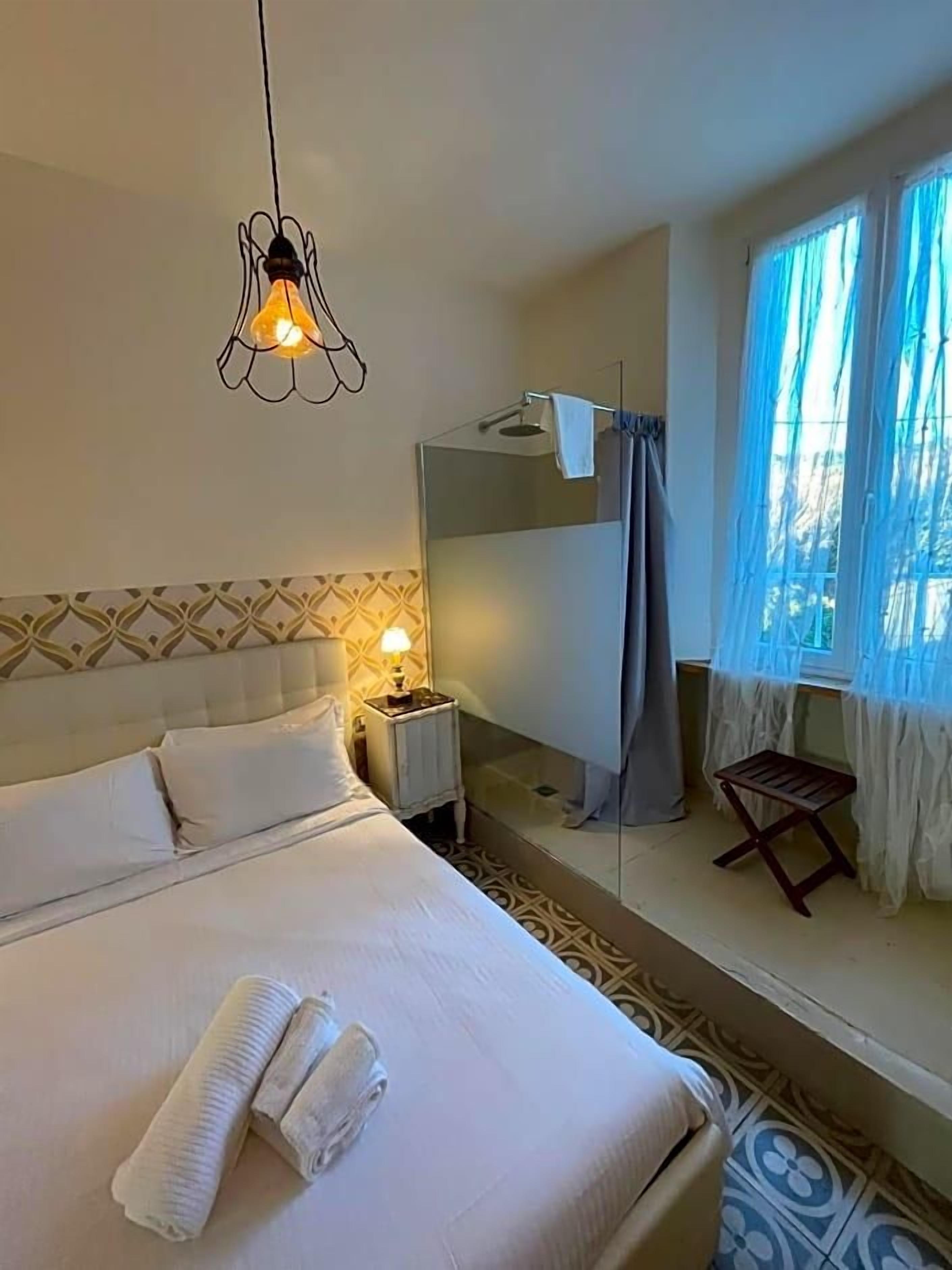 A hotel room with a bed, a bedside lamp, a small table, a chair, and a window with curtains.