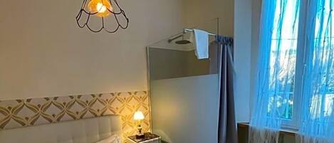 A hotel room with a bed, a bedside lamp, a small table, a chair, and a window with curtains.