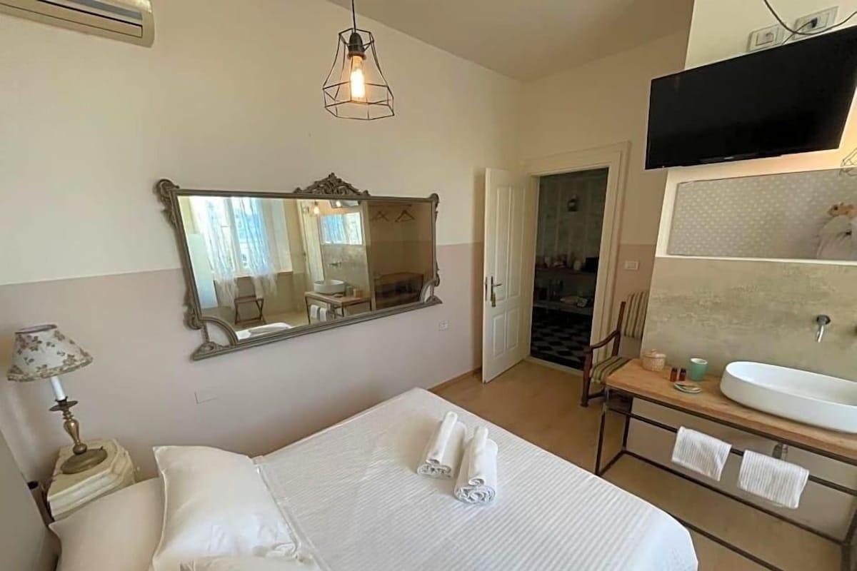 A hotel room with a bed, a large mirror, a television, a bedside table, and a bathroom with a sink and mirror.