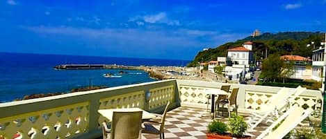 A terrace with checkered flooring, outdoor seating, and a view of the sea and buildings.
