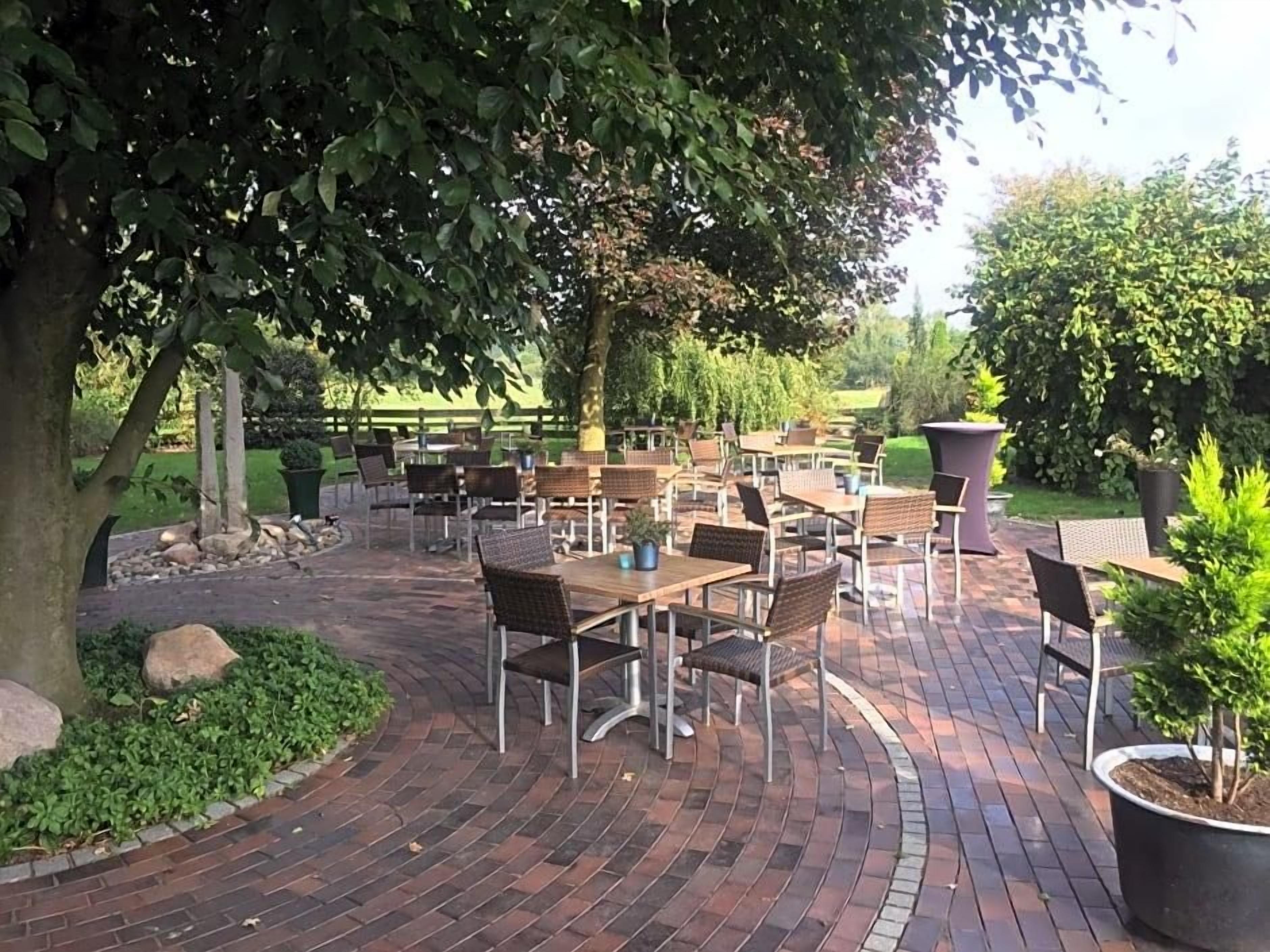 An outdoor patio area with brick flooring, multiple tables and chairs, and a large tree providing shade.