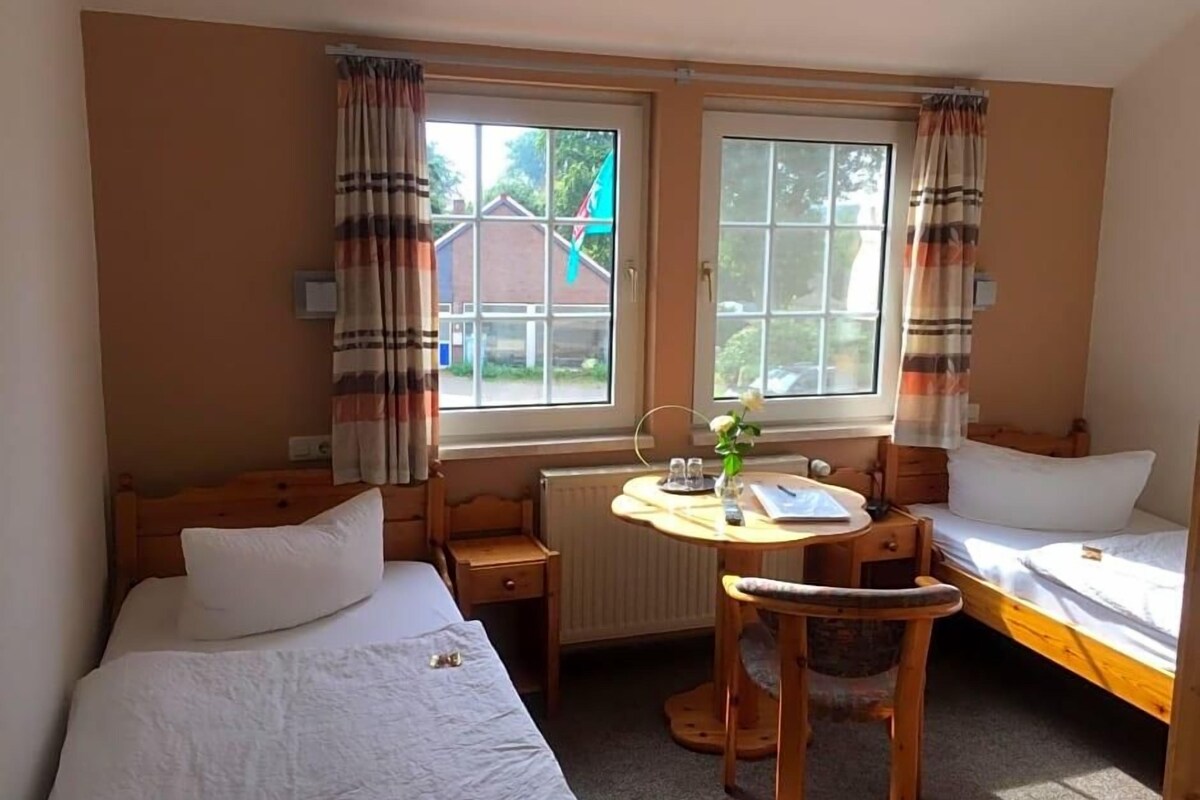 A hotel room with two single beds, a small table with a chair, a radiator, and a window with curtains.