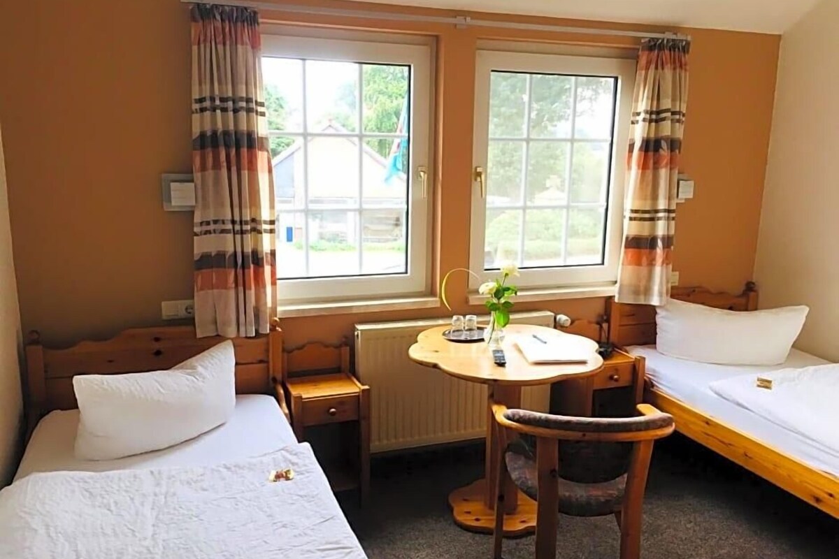 Hotellihuone, jossa on kaksi sänkyä, pieni pöytä, tuoli ja ikkuna, jossa on verhot.