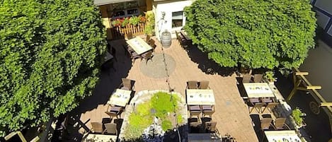 An outdoor patio area with tables and chairs arranged around a central fire pit, surrounded by lush greenery.