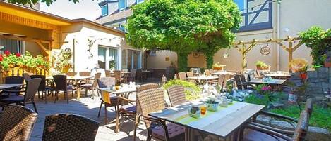 Un espace extérieur aménagé en terrasse, avec des meubles en osier, une table dressée pour les repas et un grand arbre qui offre de l’ombre.