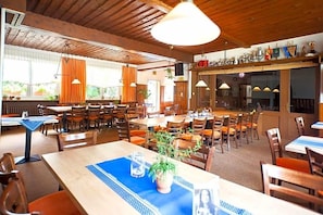A restaurant with multiple tables and chairs, wooden ceiling beams, and hanging pendant lights. - Hotel & Restaurant Ernst (Giesen)