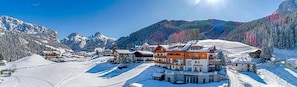 A snow-covered mountain resort with multiple buildings, including a prominent red-roofed structure, surrounded by snow-covered slopes and trees. - Hotel Chalet Dlaces (Selva di Val Gardena)