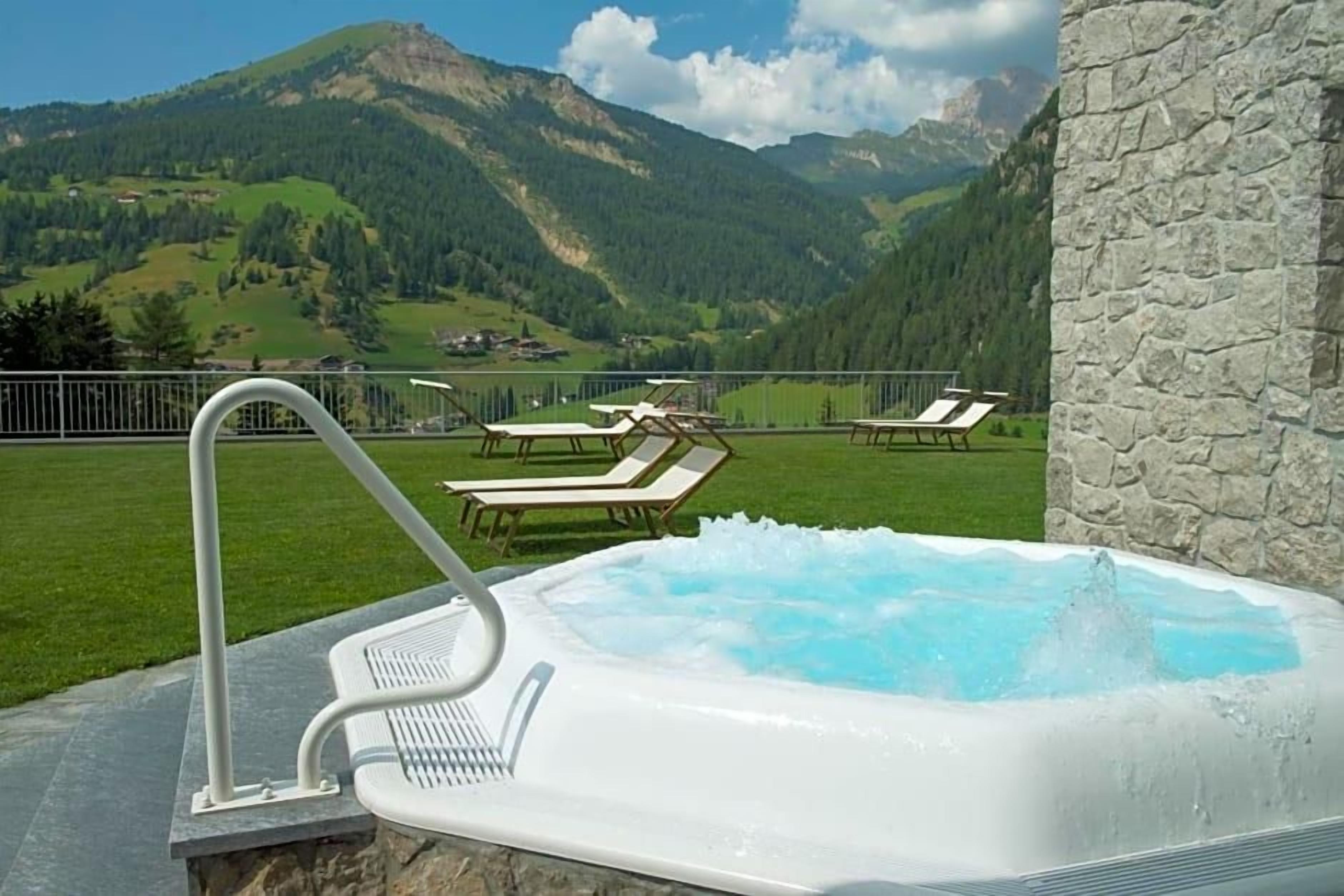 A hot tub with a mountain view, surrounded by lounge chairs and a stone wall.