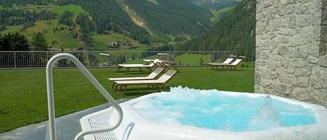 A hot tub with a mountain view, surrounded by lounge chairs and a stone wall.